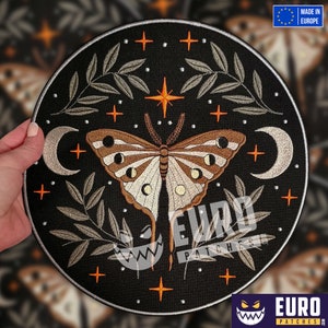 Peut inclure: Patch brodé représentant un papillon blanc et brun avec des phases lunaires sur ses ailes. Le papillon est entouré de feuilles vertes et d'étoiles orange sur un fond noir. Le patch est circulaire et possède une bordure argentée.