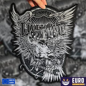 Pode incluir: Patch bordado a preto e branco com um desenho de águia e o texto "LIVE TO RIDE". O patch tem a forma de um escudo e é segurado por uma mão. A águia tem as asas abertas.