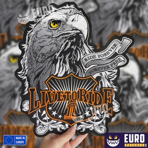 Op de afbeelding: Zwart-witte geborduurde patch met een kop van een Amerikaanse zeearend, een banner met de tekst "Ride Forever, Forever Free" en de woorden "Live to Ride" in oranje letters. De patch heeft een zwarte achtergrond met witte en oranje accenten.