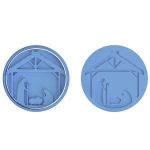 Op de afbeelding: Twee ronde blauwe ornamenten met een witte omtrek, die elk een kerststal voorstellen met een kribbe, een ster en drie figuren.