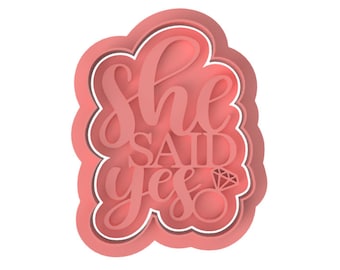 May include: Pink silicone cookie cutter with a white outline. The cutter is shaped like a heart and features the text "She Said Yes" with a diamond ring graphic.