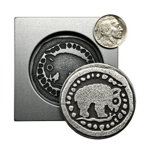 May include: A silver metal coin with a detailed design of a bison in a circle. The coin is sitting on a metal surface with a similar design of a bison in a circle.