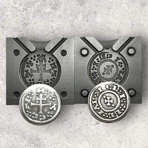 May include: Two metal molds for making coins, with a silver coin resting on each mold. The molds are gray and have a textured surface. The coins are silver and have a cross design with a circle of dots around the edge.