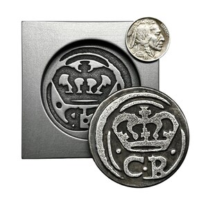 May include: A silver metal coin mold with a raised design of a crown and the letters "C.R." The mold is sitting on a silver metal surface. A silver coin with a similar design is sitting next to the mold.