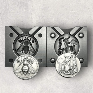 May include: Two silver metal molds for casting coins. The left mold has a bee design and the right mold has a deer design. A silver coin with a bee design is sitting on the left mold and a silver coin with a deer design is sitting on the right mold.