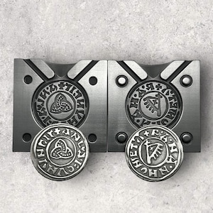 May include: Two metal molds for making round metal pendants. The molds are made of silver metal and have a black design. The design on the left mold is a Celtic knot, and the design on the right mold is a Viking rune.