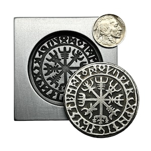 May include: A silver coin with a black and white design of a compass with runes around the edge. The coin is sitting on a metal mold with the same design.