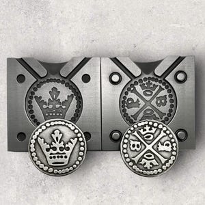 May include: Two silver metal molds for making coins. One mold has a crown design, the other has a cross with crowns at each end.  The molds are next to each other on a gray surface.