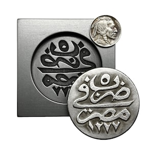 May include: A silver coin with Arabic calligraphy in a raised design. The coin is next to a metal mold with the same design.