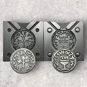 May include: Two metal molds for making coins. The molds are made of a dark gray metal and have a circular design. The left mold features a tree with three branches and the right mold features a chalice with a stem. Both molds have Hebrew text around the edges.