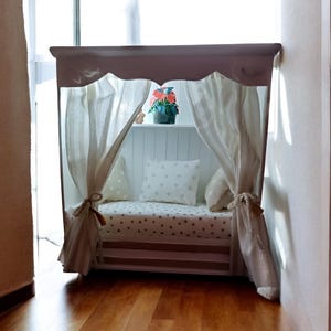 Può includere: Un letto per bambini bianco e marrone chiaro con baldacchino. Il letto ha una tenda a righe, un cuscino a forma di cuore e una piccola composizione floreale. Il letto è posto davanti a una finestra.
