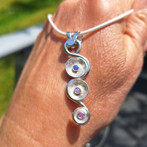 Könnte beinhalten: Eine silberne Anhänger Halskette mit einem Spiraldesign. Der Anhänger hat drei kreisförmige Elemente, jedes mit einem kleinen, farbigen Edelstein. Die Halskette hat ein weißes Band.