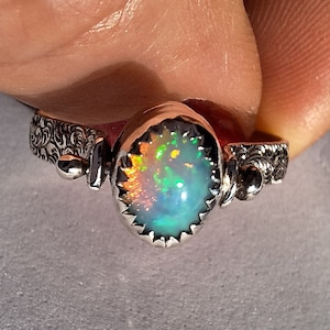 Könnte beinhalten: Silberring mit einem ovalen Opal-Edelstein, der eine lebendige Farbpalette zeigt. Der Opal ist in einer Lünettenfassung mit gezacktem Rand gefasst. Der Ring hat ein verziertes, graviertes Design.