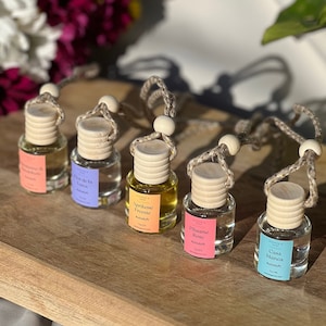 Peut inclure: Six petites bouteilles en verre avec des bouchons en bois et des cordons suspendus. Les bouteilles sont remplies d'un liquide clair et portent des étiquettes avec différents parfums : "Apricot Freesia", "Prune Rose", "Casa Blanca", "Verde de la Luna", "Vanilla" et "Mandarin".