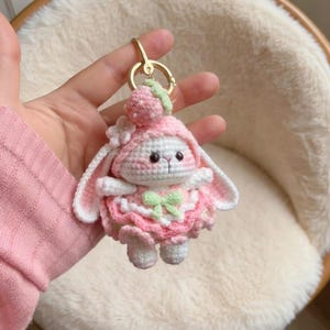 May include: A handmade crochet bunny keychain in pink and white. The bunny wears a pink dress with a green bow and a matching hat with a strawberry on top. The keychain has a gold-colored ring.