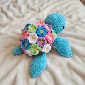 Peut inclure: Une tortue bleue au crochet faite à la main, avec une carapace ornée de fleurs au crochet colorées dans les tons de rose, blanc, bleu, vert et violet. La tortue a un aspect texturé et moelleux, sur un fond blanc cassé.