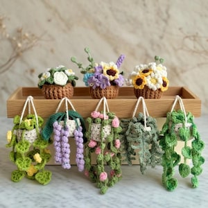 May include: Crocheted miniature plants, including hanging baskets and potted flowers, in various colors. The collection features detailed crochet work, with white, yellow, purple, and green hues. The plants are displayed on a wooden tray and hanging from white cords.