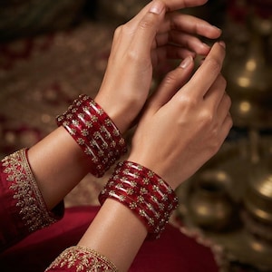 May include: A set of translucent deep red bangles featuring embedded gold-toned floral accents. The circular bracelets are worn in stacks on each wrist, showcasing a smooth, glossy finish and intricate metallic detailing throughout the red material.
