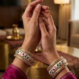 May include: A set of two gold-toned metal bangles featuring a white enamel base with intricate floral patterns in red, green, and pink. Each bangle is bordered by thin, textured gold bands, creating a layered appearance on the wrist.