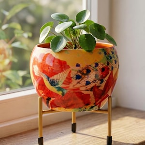 Könnte beinhalten: Ein runder Keramikübertopf mit einer leuchtend orangefarbenen und gelben Oberfläche, die mit einem bunten Kolibri- und Blumenmuster verziert ist. Der Übertopf steht auf einem goldfarbenen Metallständer mit drei Beinen und schwarzen Schutzkappen.