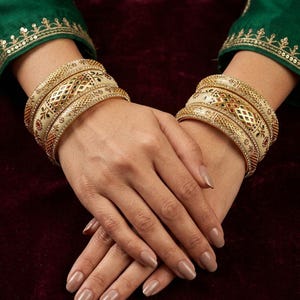 May include: A set of cream-colored bangles featuring intricate gold-tone metal filigree, small embedded red and green stones, and delicate floral patterns. These circular bracelets are worn in stacks on both wrists, showcasing a polished finish.