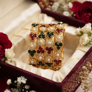 May include: A set of four gold-toned bangles featuring alternating heart-shaped red and green stones, accented with small white pearls and clear crystals. The bangles have a textured metal finish and are arranged in a vertical stack.