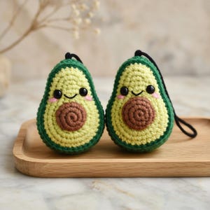 May include: Two crocheted avocado ornaments with smiling faces and black eyes. The avocados are green with a light green center and a brown seed. Each ornament has a black loop for hanging. They are displayed on a wooden tray.