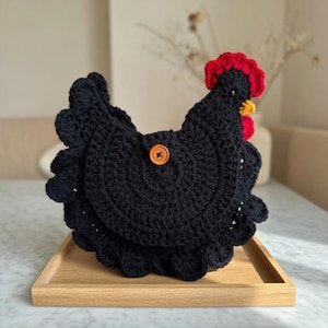 Könnte beinhalten: Handgefertigte, schwarze Häkeltasche in Hühnerform mit rotem Kamm und gelbem Schnabel. Die Tasche hat einen Knopfverschluss und einen gewellten Rand. Sie liegt auf einem Holztablett.