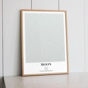 May include: A framed print with a light wood frame. The print features a textured, speckled gray background and the word "MOON" in a bold, sans-serif font at the bottom. Additional text is below the word "MOON".