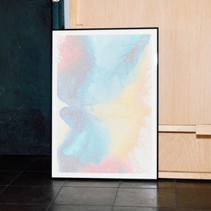 Puede incluir: Impresión artística abstracta en un marco negro. La obra de arte presenta un diseño colorido con tonos de azul, rosa y amarillo. La impresión se muestra contra una pared oscura y un panel de madera clara.
