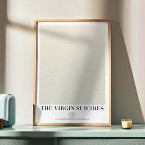 Może przedstawiać: Oprawiony plakat z tytułem "THE VIRGIN SUICIDES" w minimalistycznym designie. Plakat jest w jasnej drewnianej ramie i spoczywa na miętowej zielonej powierzchni, obok niebieskiego wazonu i drewnianego pudełka. W pobliżu stoi zapalona świeca.