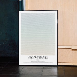 May include: A framed poster with a light blue and white gradient background. The poster has a black frame and the text "THE LORD OF THE RINGS THE TWO TOWERS" at the bottom. The poster is leaning against a wall.