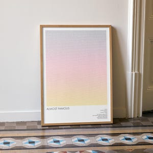 Puede incluir: Un póster enmarcado con un diseño degradado, que va del gris en la parte superior al rosa, luego al amarillo en la parte inferior. El póster se titula "Almost Famous" con texto adicional debajo. El marco es de madera marrón claro.
