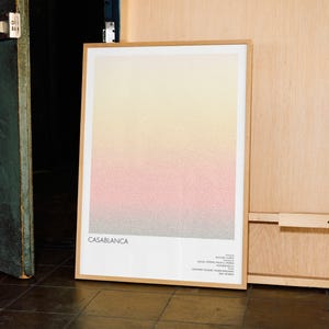 Pu&ograve; includere: Una stampa artistica incorniciata con un design sfumato in giallo, rosa e grigio. La parola "CASABLANCA" &egrave; stampata in basso, insieme a testo aggiuntivo. La stampa &egrave; in una cornice di legno chiaro, appoggiata a una parete di legno.