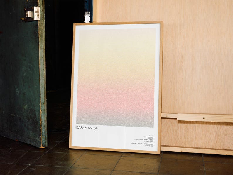 Pu&ograve; includere: Una stampa artistica incorniciata con un design sfumato in giallo, rosa e grigio. La parola "CASABLANCA" &egrave; stampata in basso, insieme a testo aggiuntivo. La stampa &egrave; in una cornice di legno chiaro, appoggiata a una parete di legno.