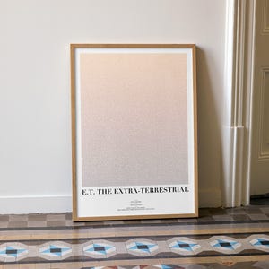 Puede incluir: Un póster enmarcado con el texto "E.T. THE EXTRA-TERRESTRIAL" en la parte inferior. La obra de arte tiene un fondo degradado de color rosa claro y gris. El póster está apoyado contra una pared blanca.