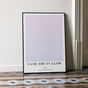 May include: A framed art print with a gradient background of pink and lavender hues. The print features the text "I SAW THE TV GLOW" in a minimalist design. The artwork is leaning against a white wall.