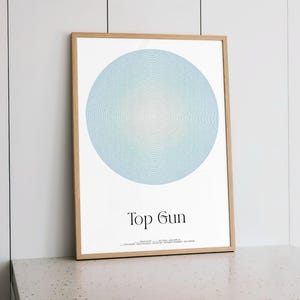 Puede incluir: Un póster enmarcado con un diseño circular azul claro con efecto degradado. El póster se titula "Top Gun" en una fuente moderna. La obra de arte se muestra en un marco de madera clara, ideal para la decoración.