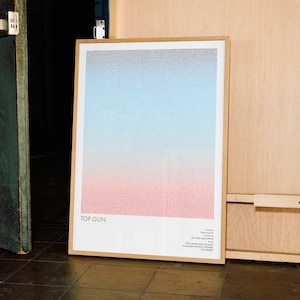 May include: A framed poster with a gradient design, transitioning from pink at the bottom to blue at the top. The poster is titled "TOP GUN" and includes text at the bottom. The frame is a light brown color.
