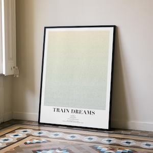 May include: A framed print with the title "TRAIN DREAMS" in a black frame. The artwork displays a colour gradient, shifting from yellow to green. The print is propped against a white wall, with a patterned tile floor visible in the foreground.