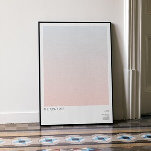 May include: Framed art print with a gradient design, transitioning from light blue to pink. The print is titled "THE GRADUATE" in a minimalist style, leaning against a white wall. The frame is black.