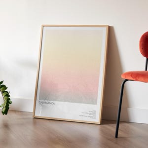 Pu&ograve; includere: Una stampa artistica incorniciata con una sfumatura di colori, dal giallo al rosa al grigio, appoggiata a una parete bianca. La stampa &egrave; etichettata "Casablanca" in basso. Una sedia rossa con gambe nere &egrave; a destra.