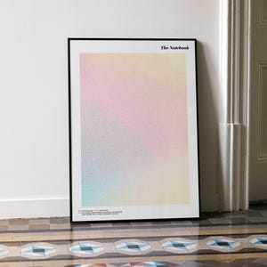 May include: A framed art print with a gradient of pastel colors, including pink, yellow, and blue. The print is titled "The Notebook" in the upper right corner. The frame is black, and the artwork leans against a white wall.