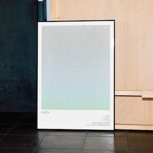 Op de afbeelding: Een ingelijste poster met een gradiëntontwerp, dat overloopt van grijs aan de bovenkant naar lichtblauw en groen aan de onderkant. Het woord "ALIEN" staat onderaan, samen met extra tekst. De poster leunt tegen een muur.