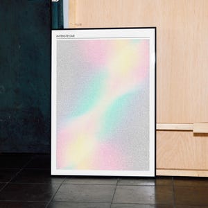 May include: Framed art print with the word "INTERSTELLAR" at the top. The artwork features a colorful, blurred design with pastel pink, yellow, and teal hues against a silver background. The print is in a black frame.
