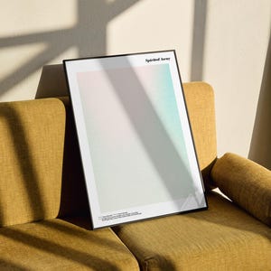 a framed poster with a minimalist design, placed on a yellow couch against a white wall.