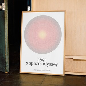 Könnte beinhalten: Gerahmter Kunstdruck mit einem großen, konzentrischen Kreisdesign in Rosa-, Grau- und Gelbtönen. Der Druck trägt den Titel "2001: a space odyssey" in schwarzer Schrift. Das Kunstwerk ist in einem hellbraunen Holzrahmen präsentiert.