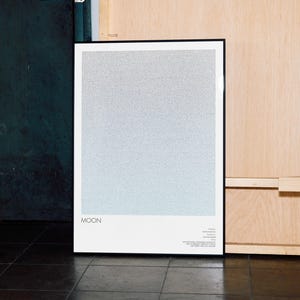 May include: A framed art print featuring a gradient design, transitioning from light blue to gray. The word "MOON" is printed in black at the bottom left, with additional text below. The frame is black, and the print is displayed against a wooden wall.
