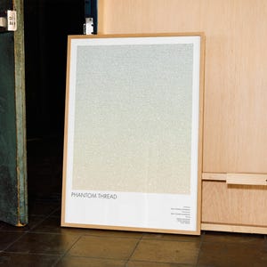 Può includere: Un poster incorniciato con il titolo "Phantom Thread" in testo bianco. Il poster presenta uno sfondo sfumato di azzurro, giallo e bianco. La cornice è in legno chiaro. Il poster è appoggiato a una parete di legno.