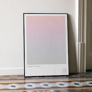 May include: A framed art print with a pink to grey gradient design. The words "UNDER THE SKIN" are printed in white at the bottom. The black framed artwork is leaning against a white wall, next to a white door.